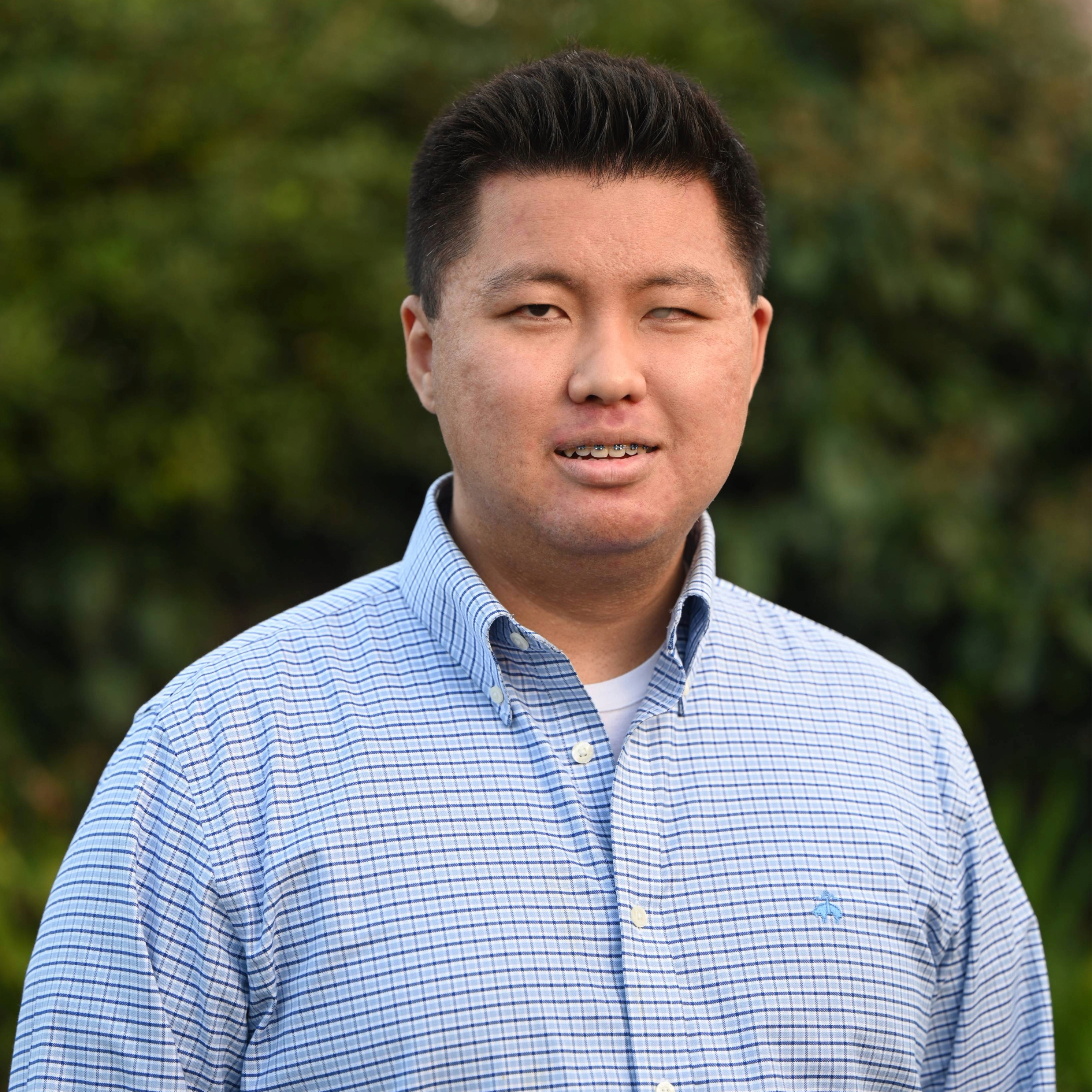 A photo of Gene, a Korean-American man smiling against a background with green trees. He has thick straight black hair that is longer on the front and shorter on the sides. He is wearing a blue collared shirt, and his dark brown eyes are smizing at the camera.