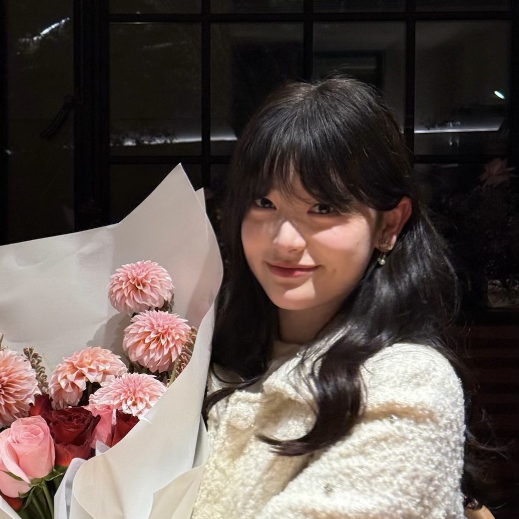Headshot of Shuqi He smiling, with long dark hair, wearing a light textured sweater, holding a bouquet of pink and red flowers indoors.